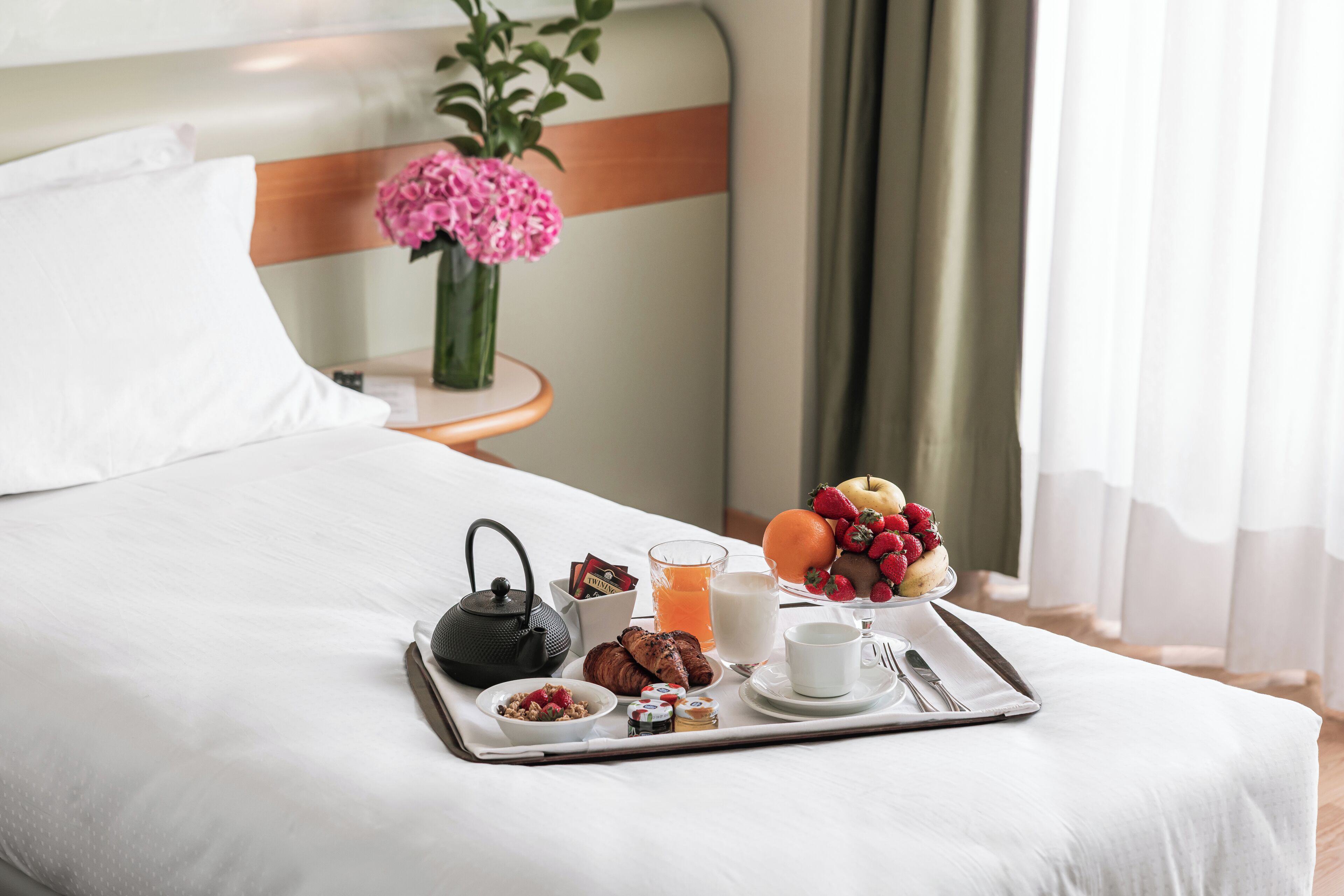 A neatly made bed with a tray of breakfast items including fruits, pastries, and beverages.