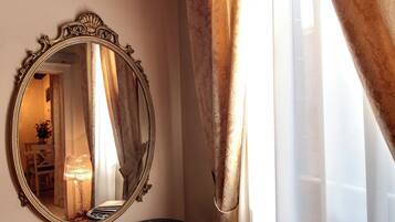 Ein verzierter Spiegel an der Wand, ein kleiner Tisch mit einer Zeitschrift und ein Fenster mit transparenten Vorhängen.