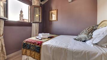 A neatly made bed with a patterned blanket, a framed picture on the wall, and a window with a view of a building.