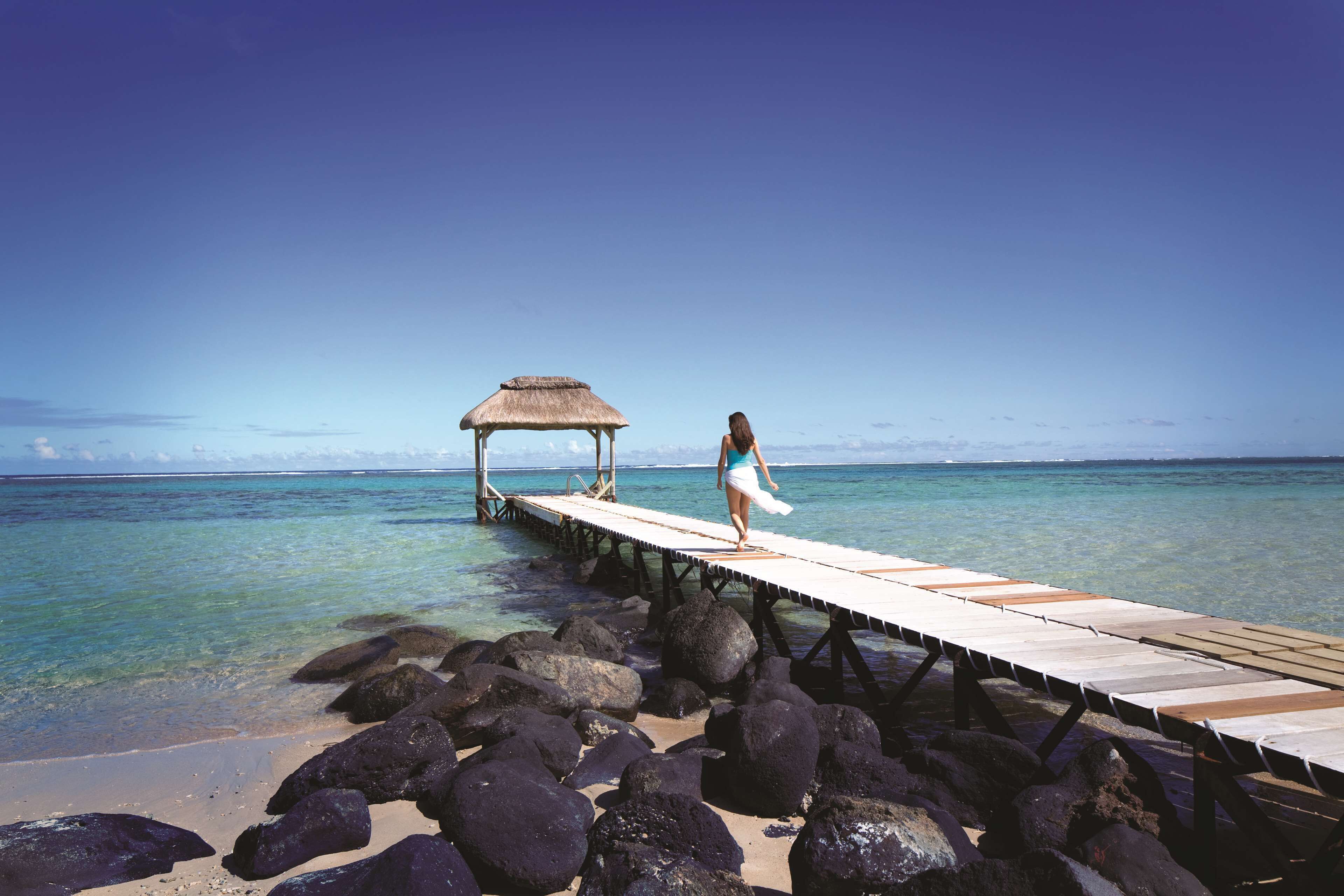 A pier extending into clear water with a thatched structure at the end and a person standing on it.