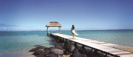 A pier extending into clear water with a thatched structure at the end and a person standing on it.