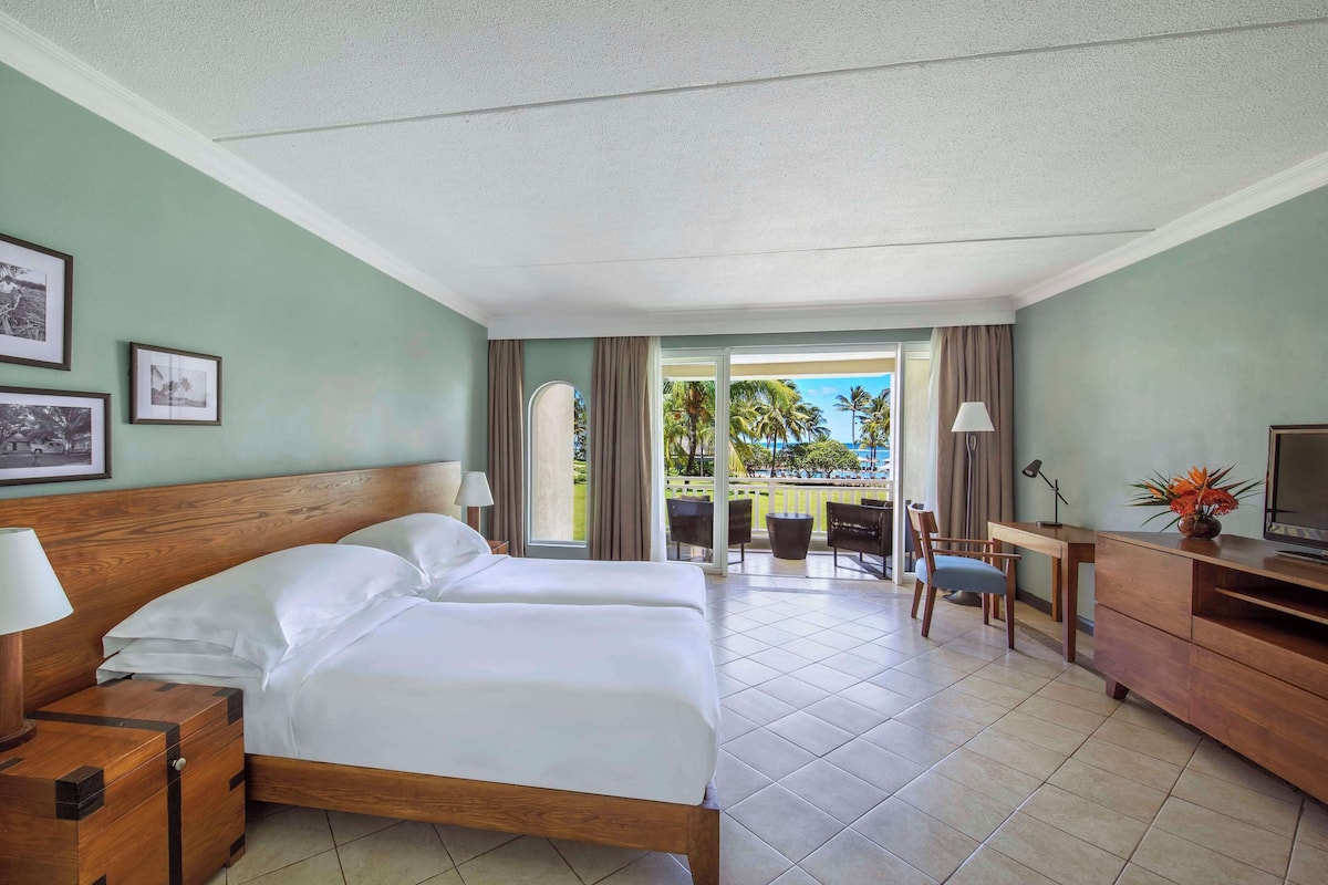 A hotel room with a bed, a desk, a TV, and a balcony with a view of palm trees.