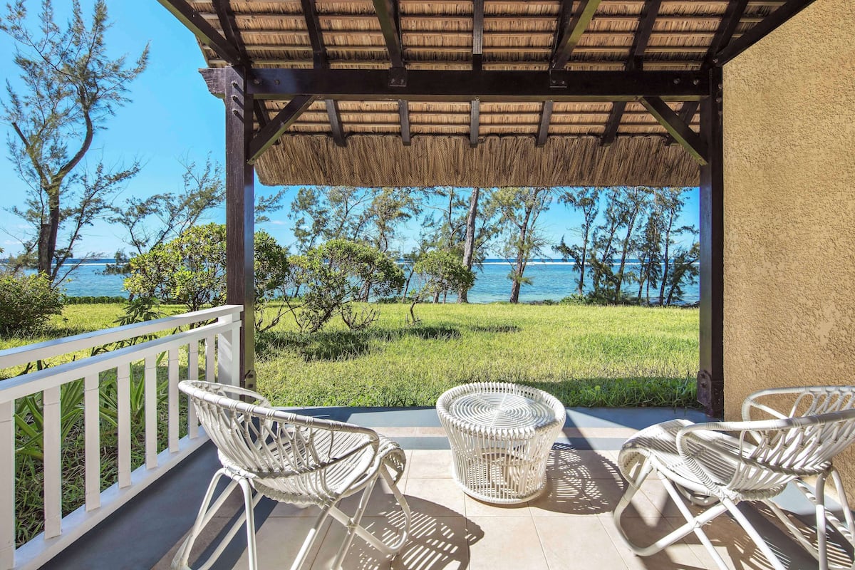 A covered outdoor seating area with wicker furniture, a view of the sea, and a grassy landscape.