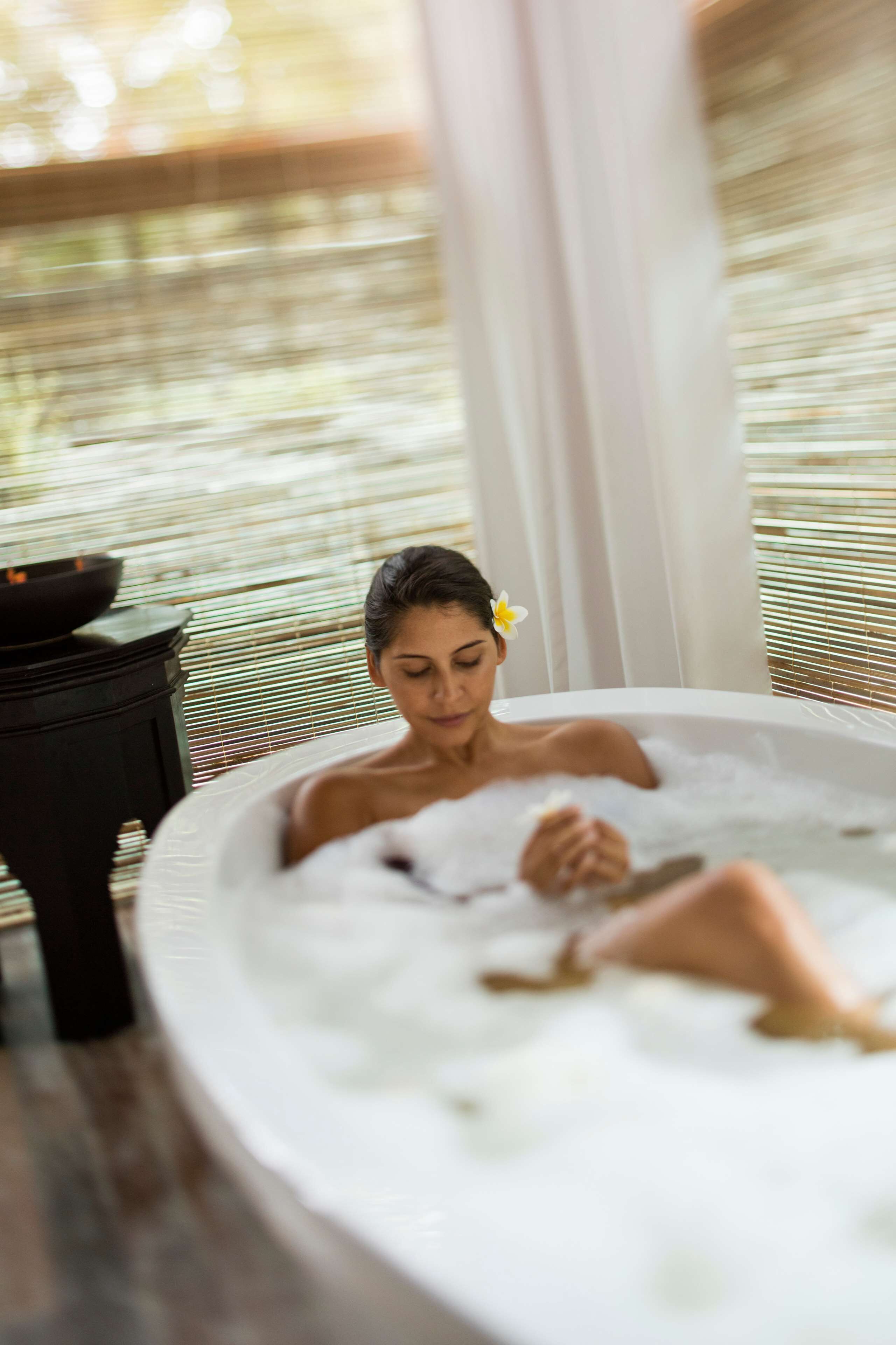 Una persona relajándose en un jacuzzi, con una flor en el cabello, una mesa negra con un bol y una ventana con persianas.