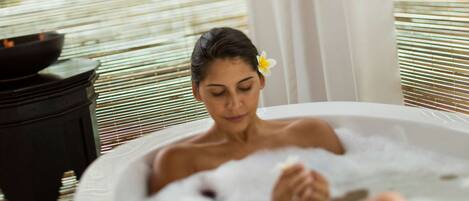 Una persona relajándose en un jacuzzi, con una flor en el cabello, una mesa negra con un bol y una ventana con persianas.