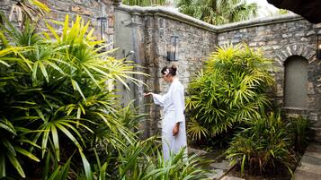 A person in a white robe standing in a lush garden with stone walls and tropical plants.