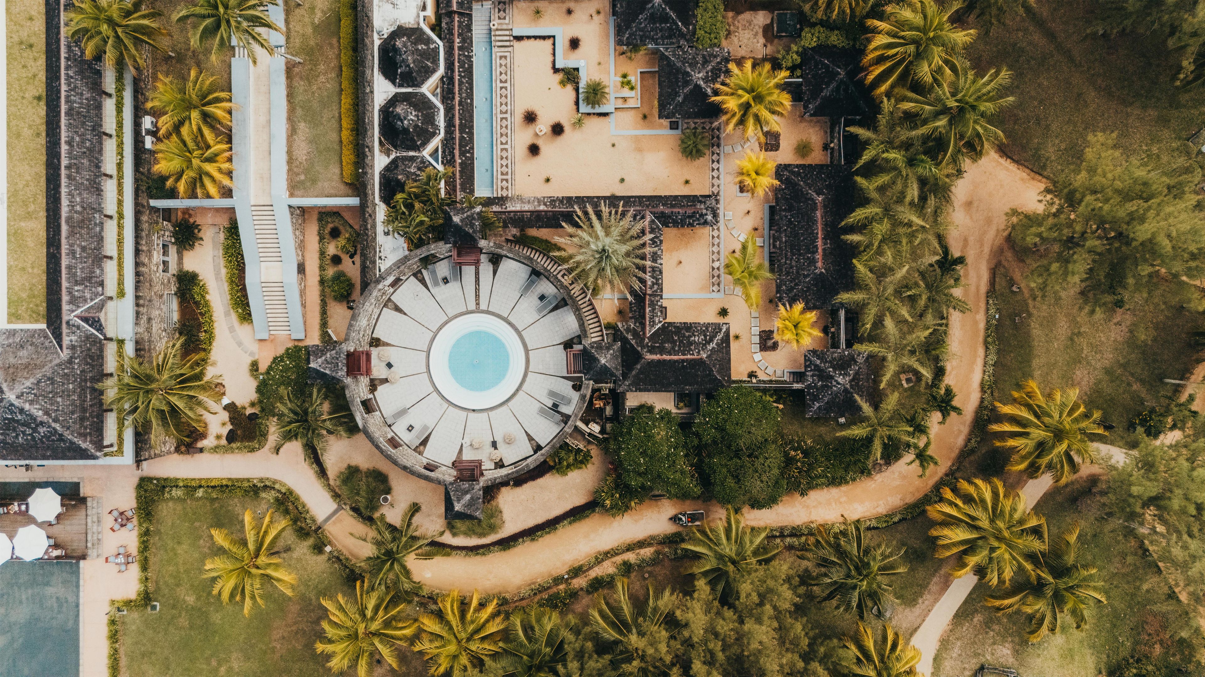 Vista aérea de un complejo turístico con una piscina circular central, rodeado de palmeras y diversos edificios.