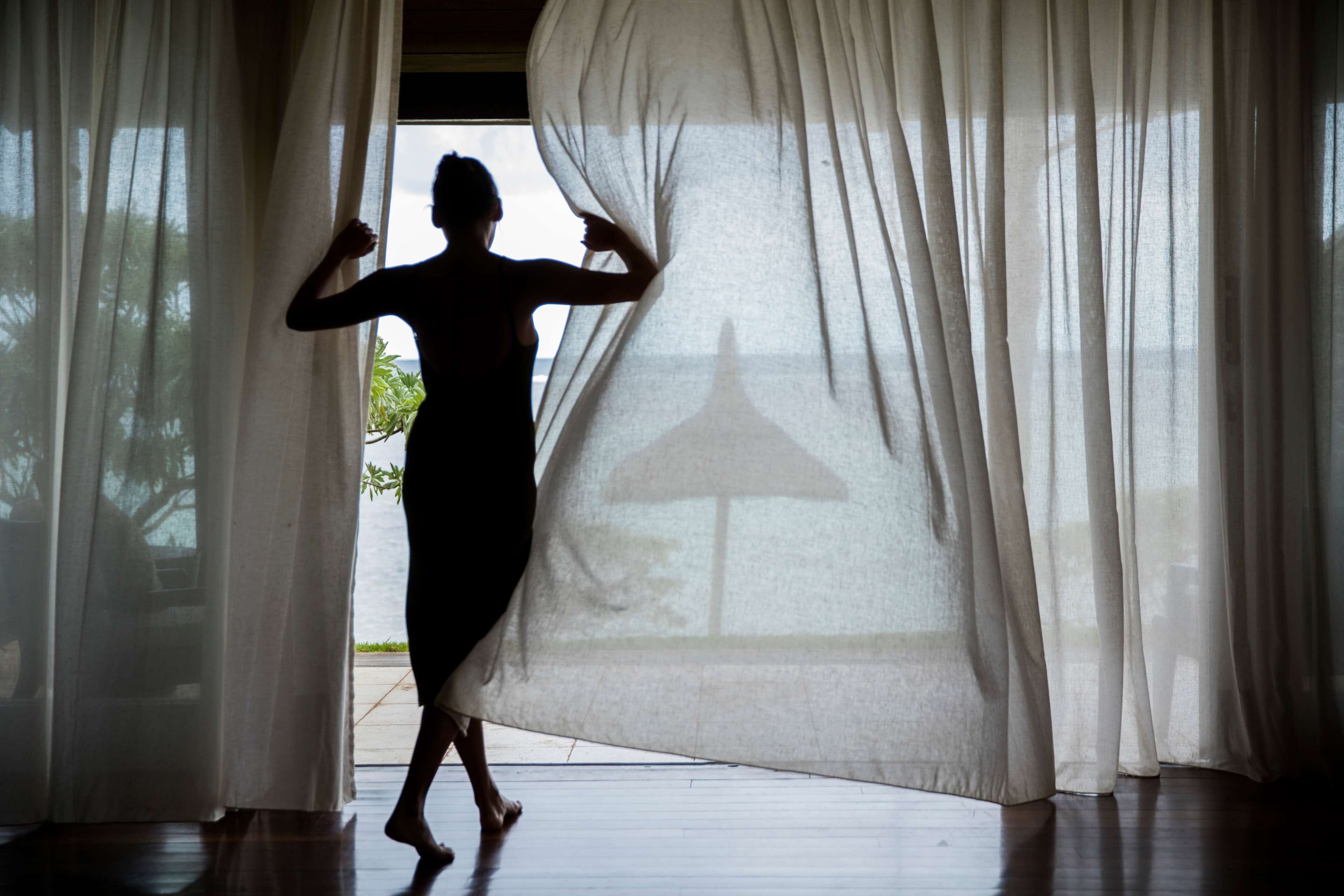 A silhouette of a person dancing in a room with sheer curtains, wooden flooring, and a view of greenery outside.