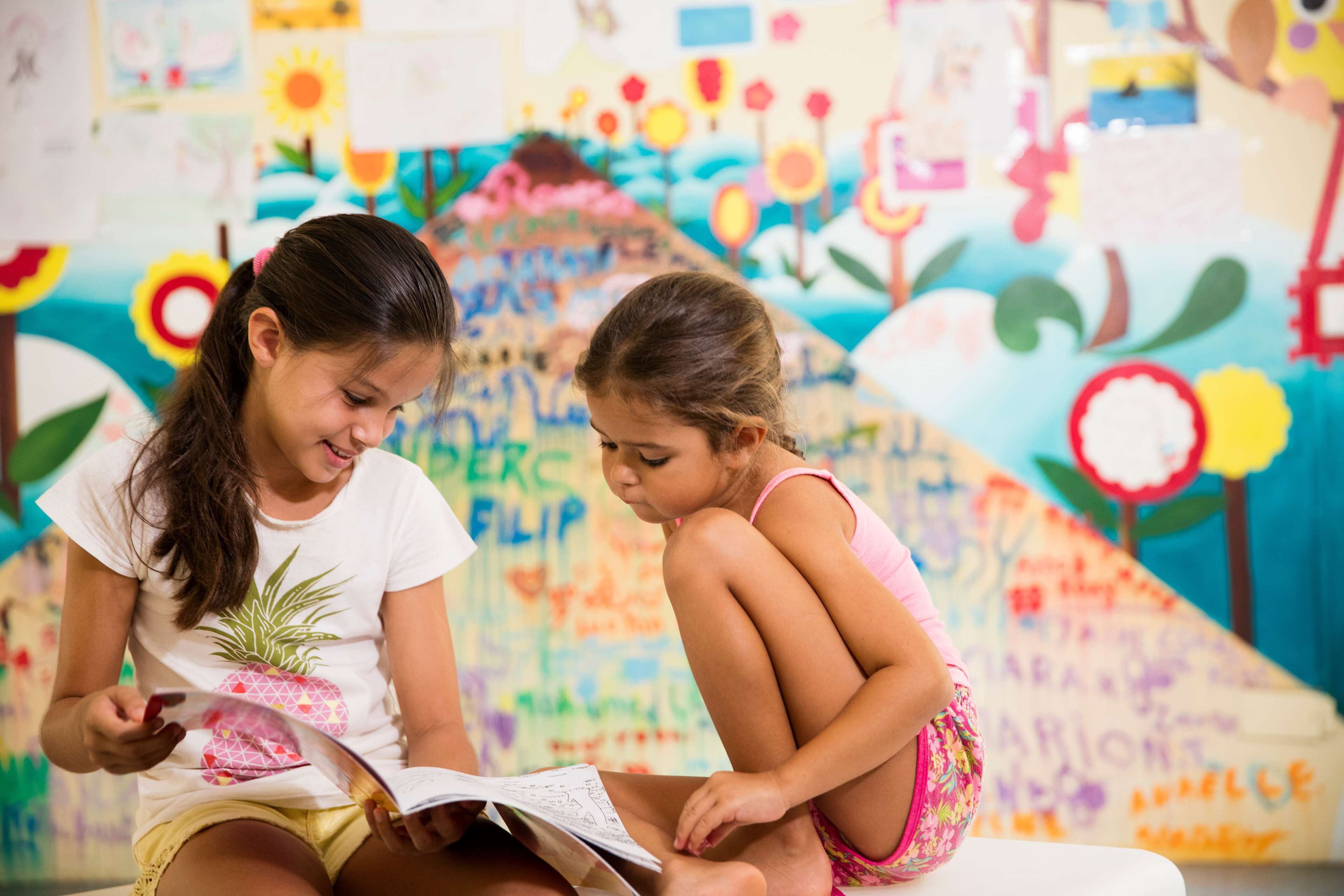 Two children reading a book in a room with colorful murals and educational decorations.