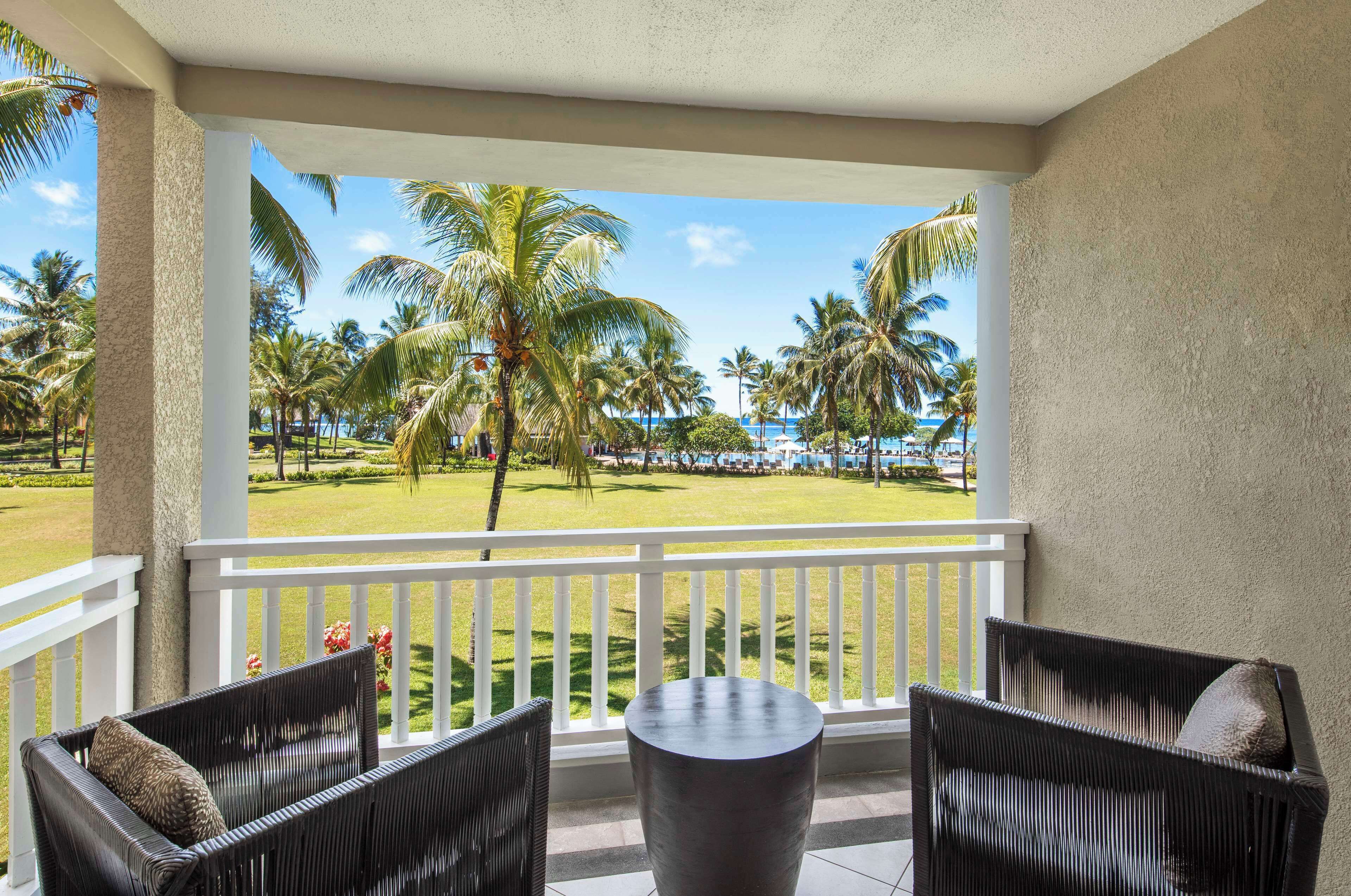 A balcony with wicker furniture, a round table, and a view of a well-maintained lawn with palm trees and a distant beach.