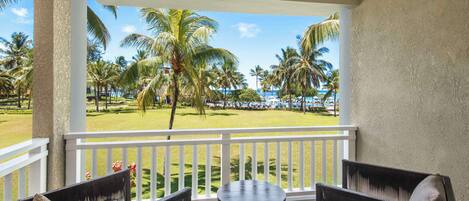A balcony with wicker furniture, a round table, and a view of a well-maintained lawn with palm trees and a distant beach.