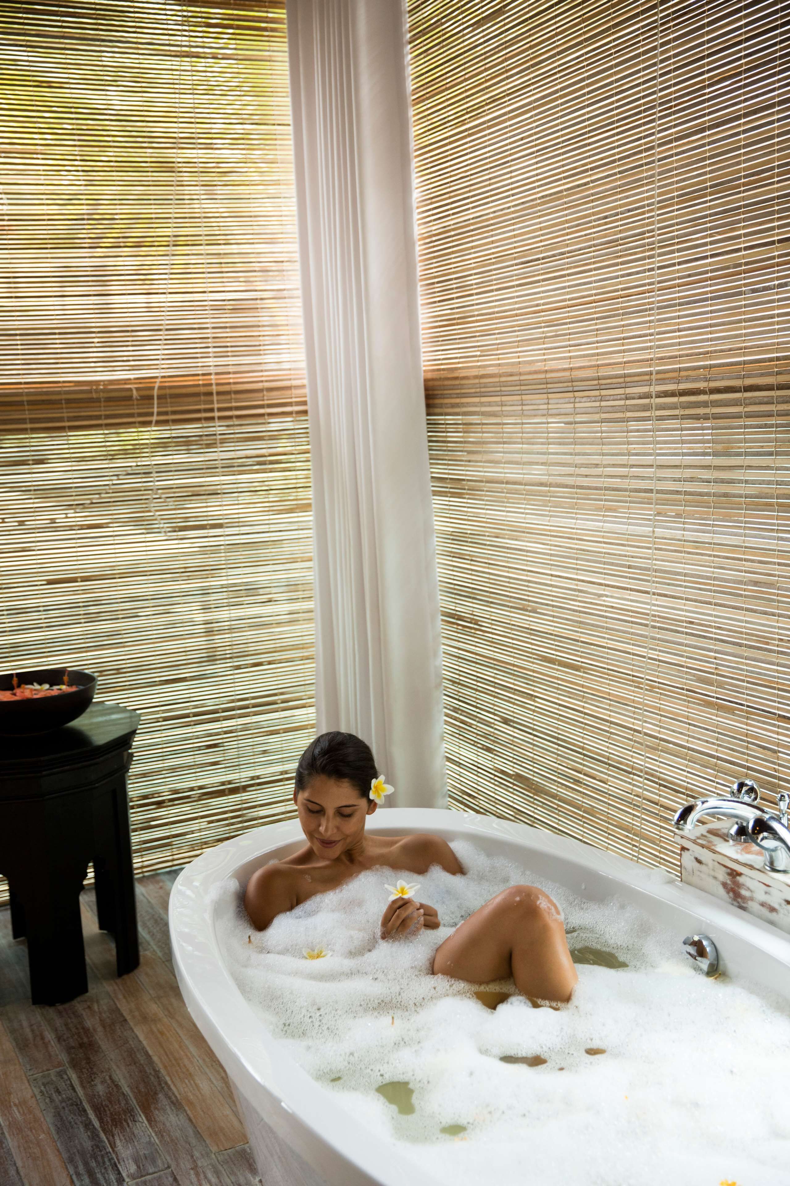 A bathtub with bubbles, a black stool, and a window with blinds.