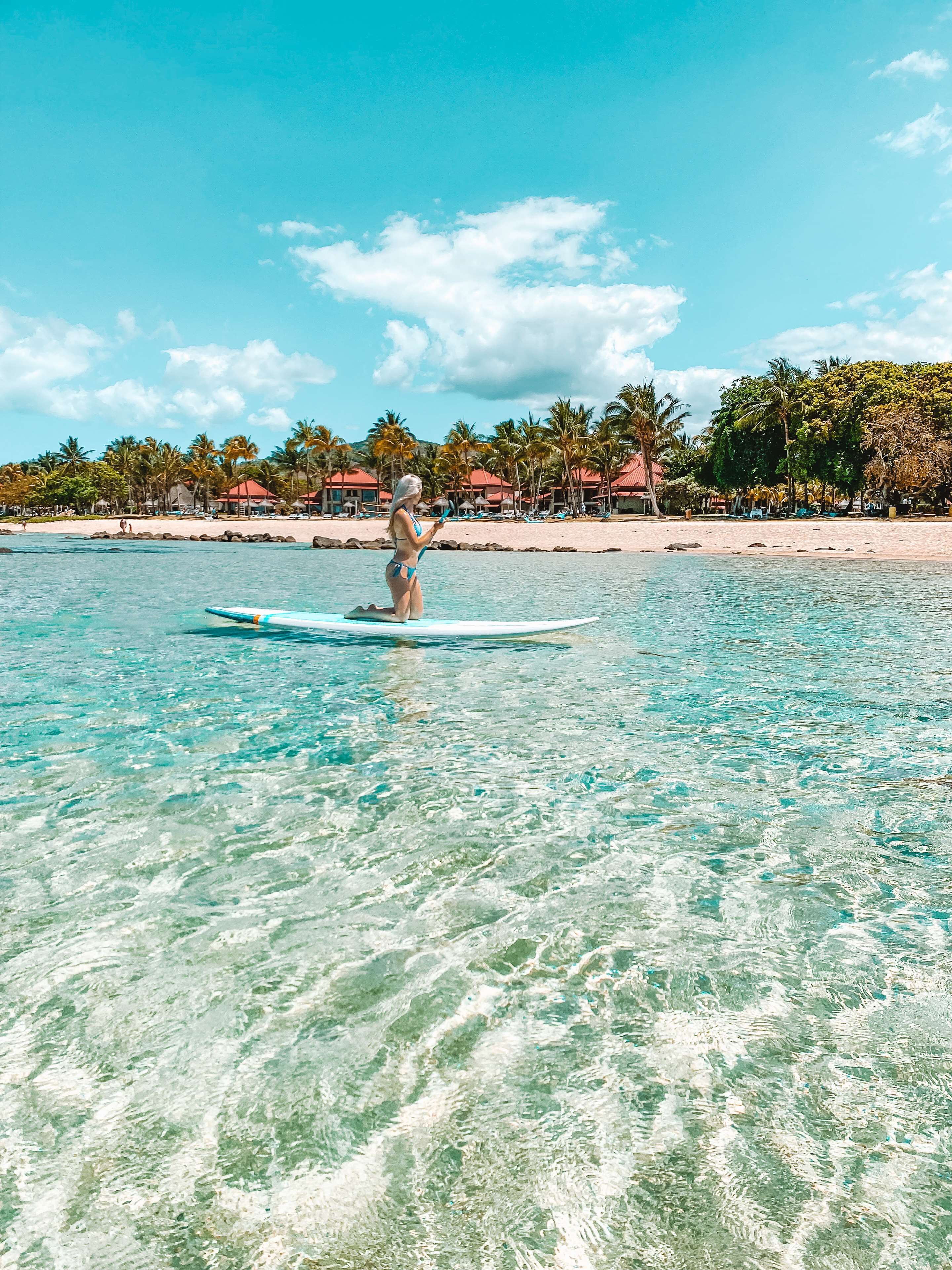 A clear tropical beach with a person paddleboarding, palm trees, and beach huts in the background.