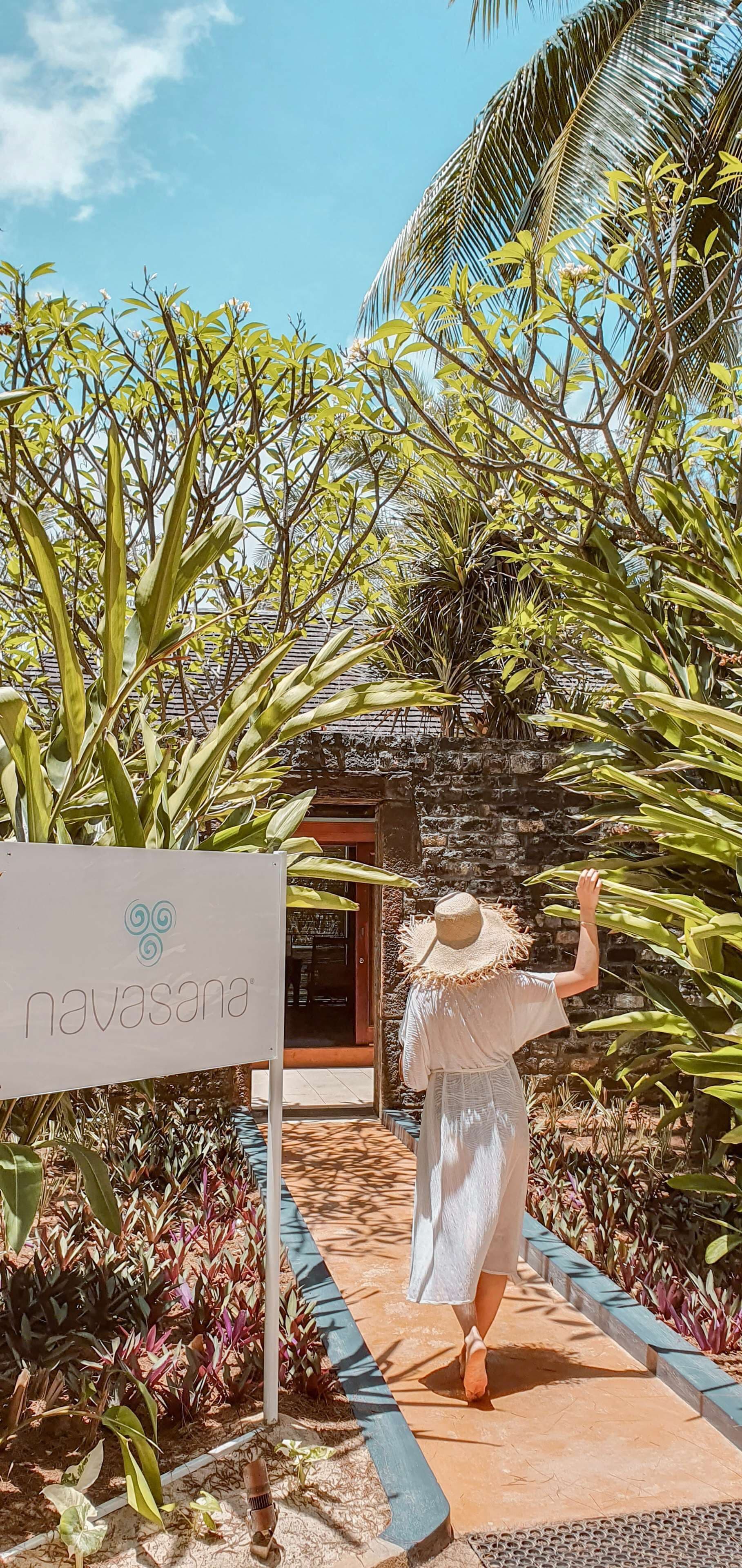 A person walking towards a building with a sign reading 'navasana' in a tropical garden setting.