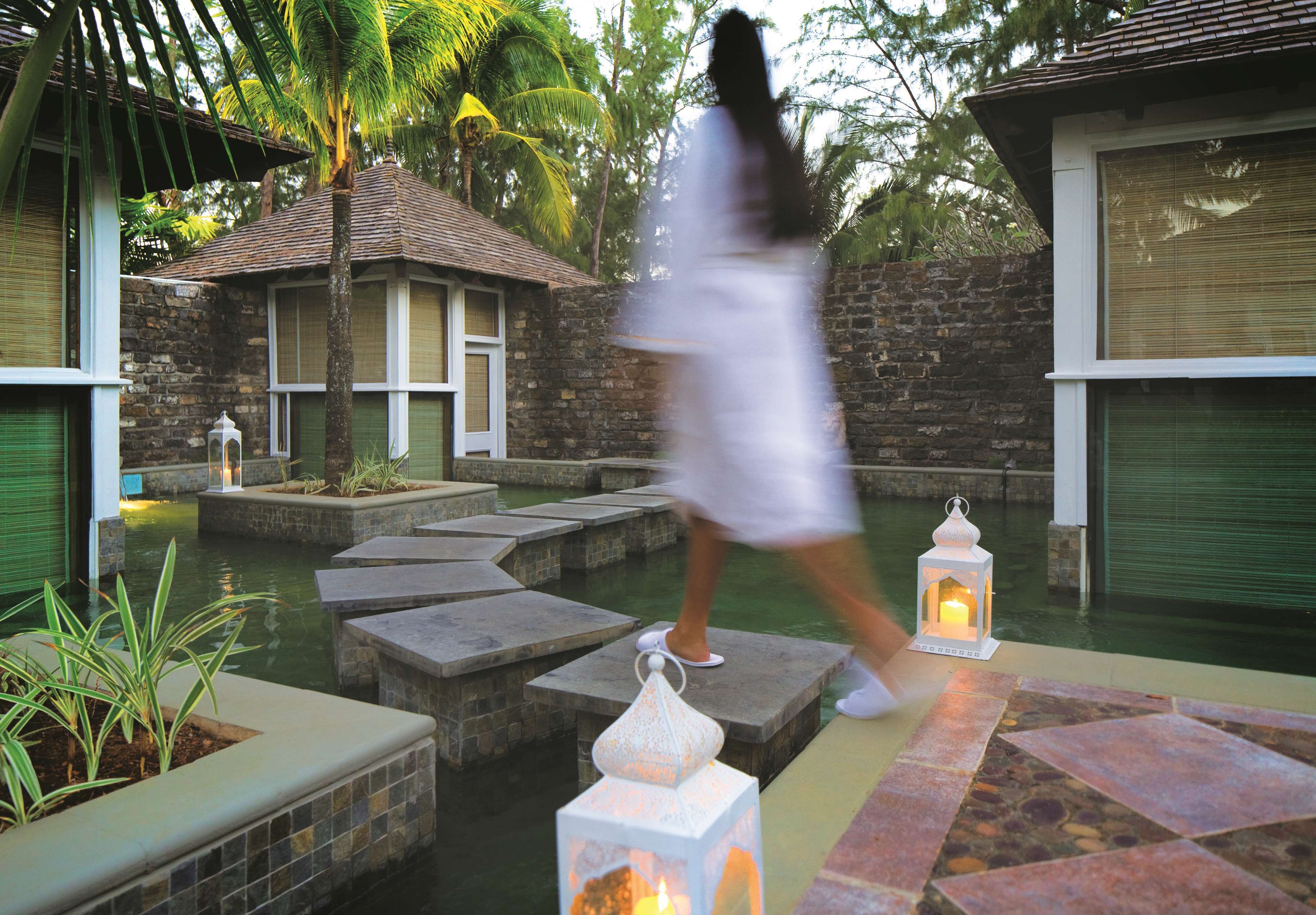 A person walking over stone steps in a courtyard with a pool, surrounded by buildings with windows and a palm tree.