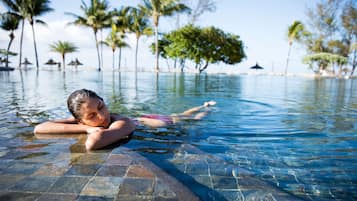 Una persona relajándose en una piscina cristalina con borde de azulejos, rodeada de palmeras y un cielo despejado.