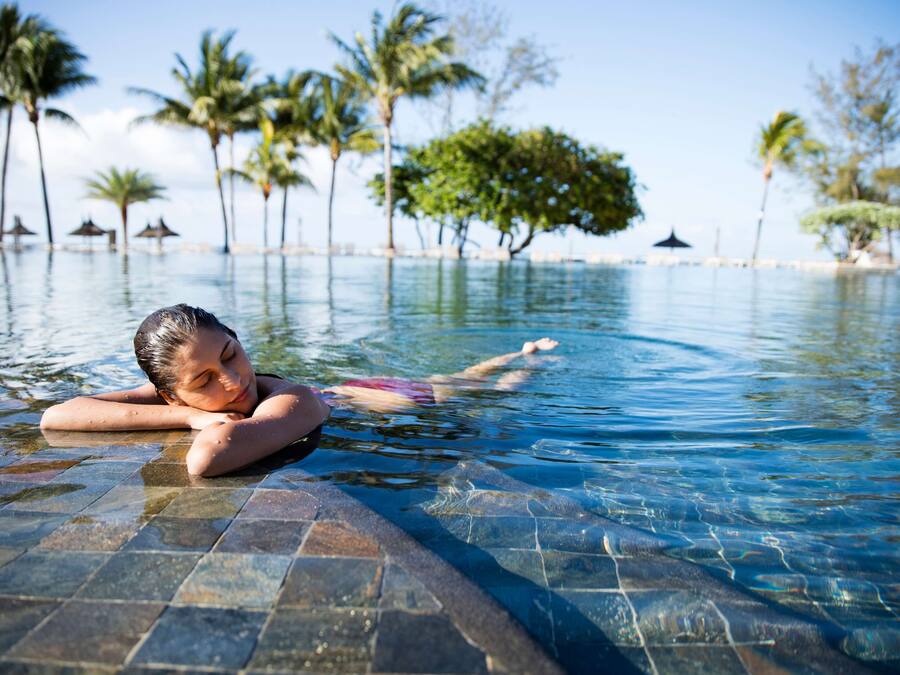 Una persona relajándose en una piscina cristalina con borde de azulejos, rodeada de palmeras y un cielo despejado.