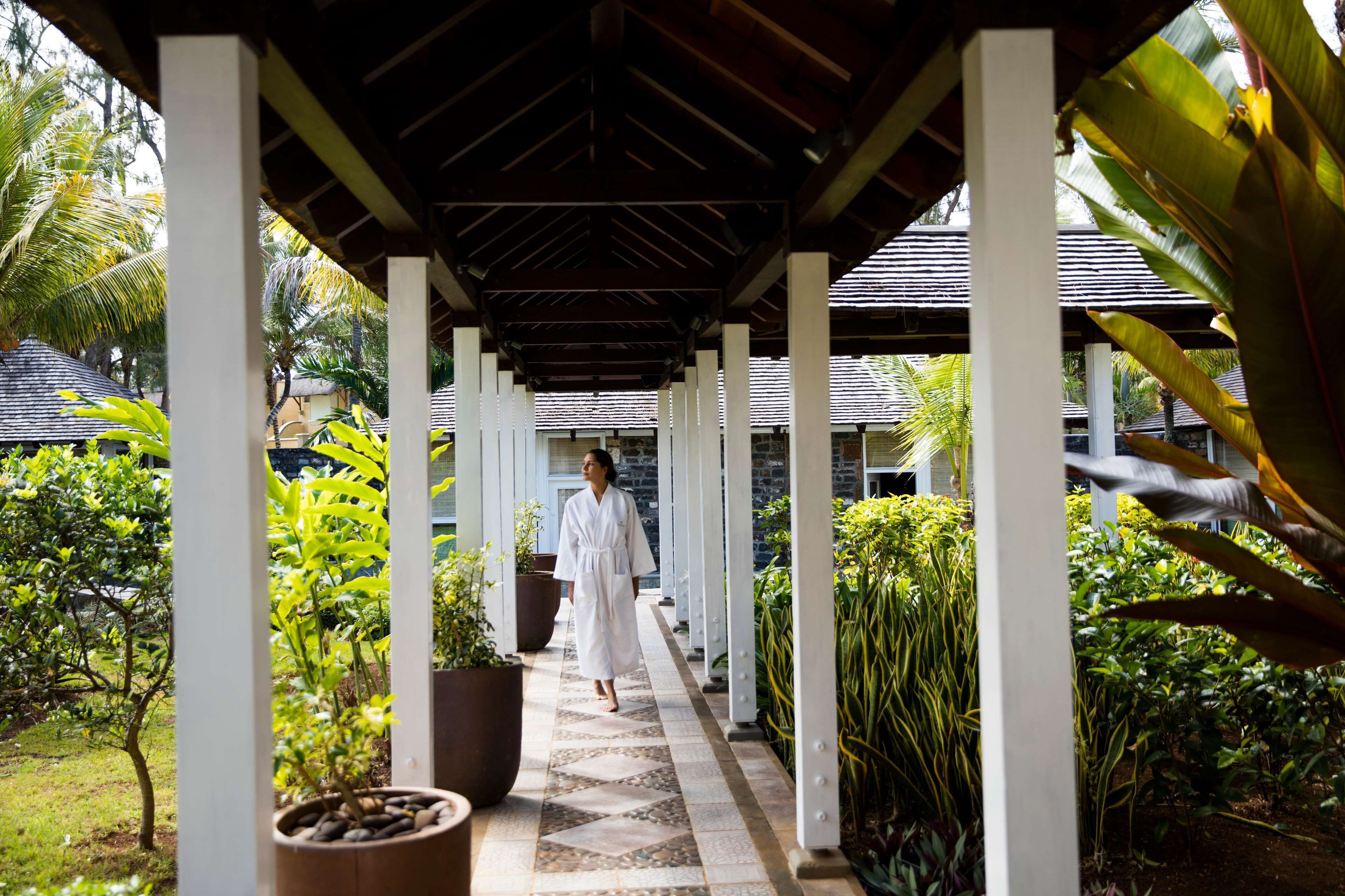 A covered walkway with a person in white, surrounded by lush greenery and potted plants.
