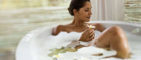 Eine Person in einer Badewanne mit Seerosen, ein Fenster mit Jalousien und ein heller Innenraum.