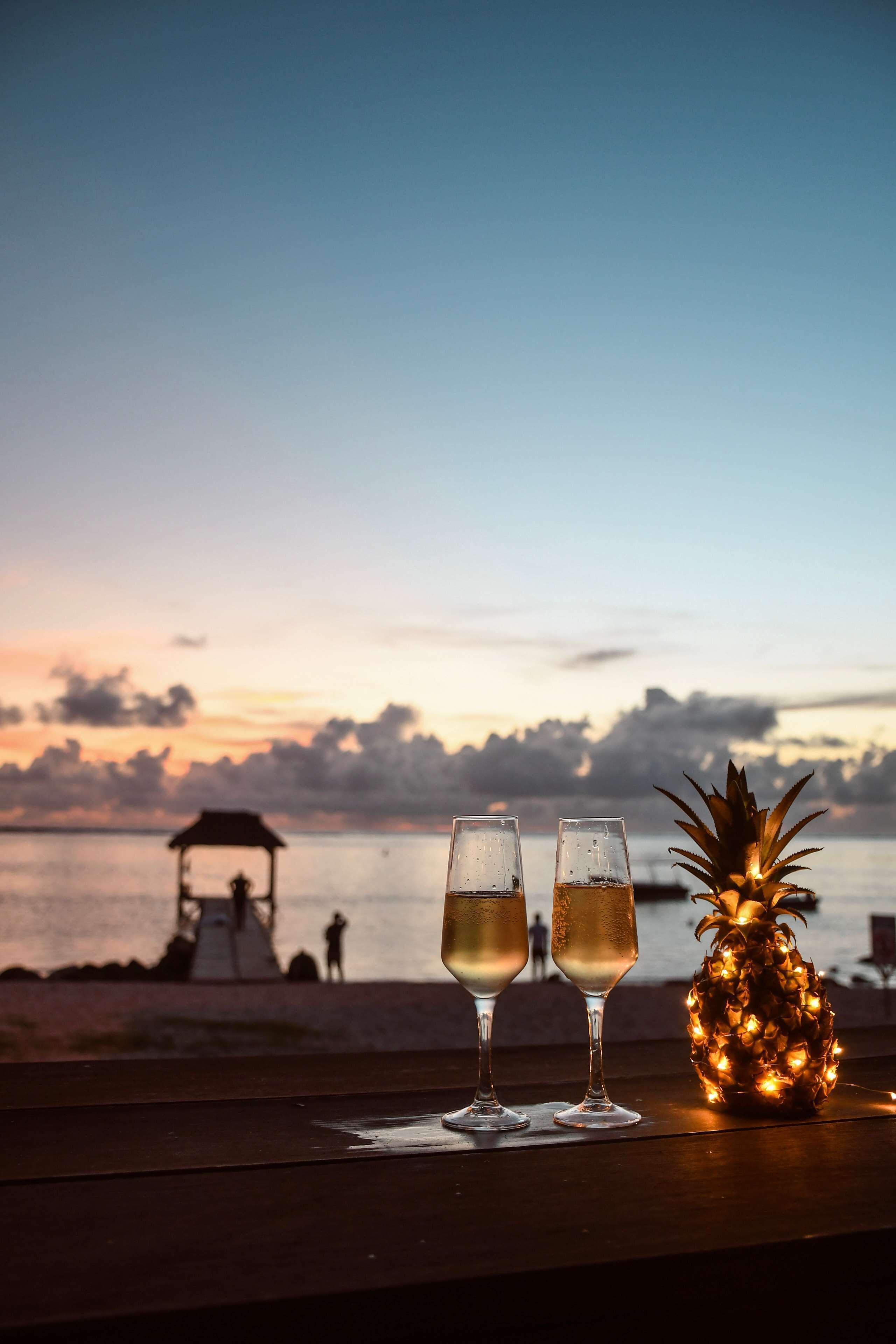 Deux verres contenant un liquide transparent sont posés sur une surface en bois. On aperçoit un ananas décoré de guirlandes lumineuses, ainsi qu’une plage avec un kiosque en arrière-plan.