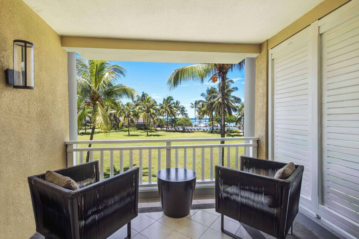 A balcony with wicker furniture, a small table, and a view of palm trees and a pool.