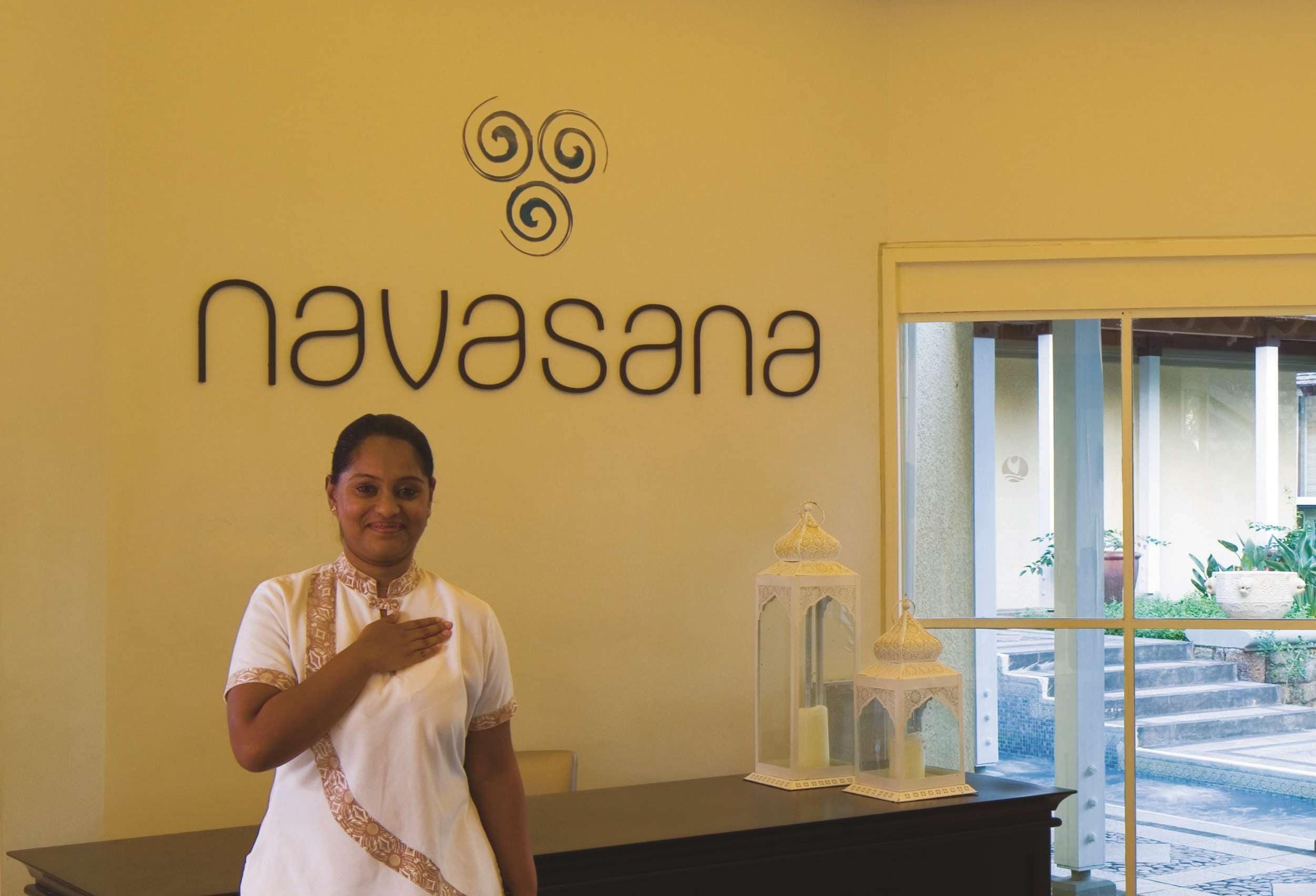 A woman standing in front of a reception desk with a sign that reads 'navasana' and a decorative lantern on the desk.