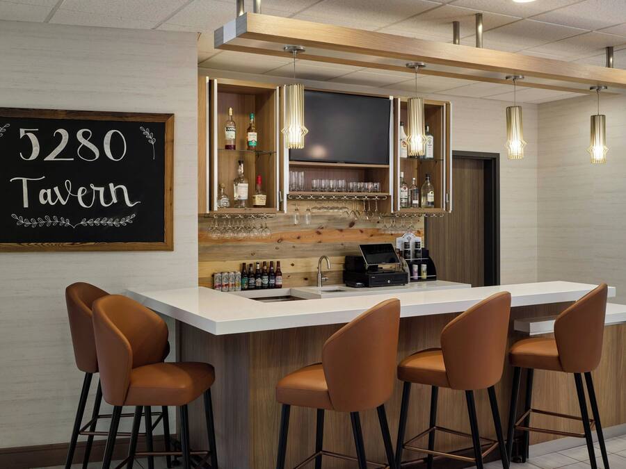 Zona de bar moderna con un letrero de pizarra que dice '5280 Tavern', taburetes, una caja registradora y una variedad de botellas y vasos.