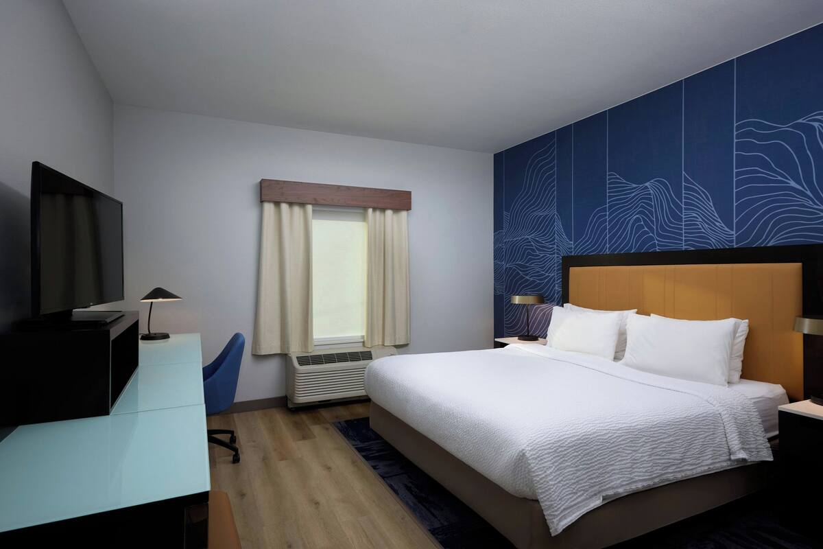 A modern hotel room with a large bed, a desk with a computer, a TV, and a window with curtains.