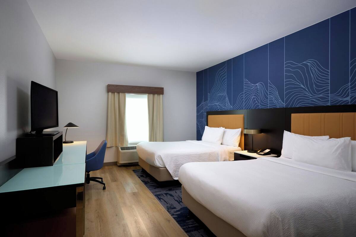 A hotel room with two beds, a desk, a TV, and a window with curtains.
