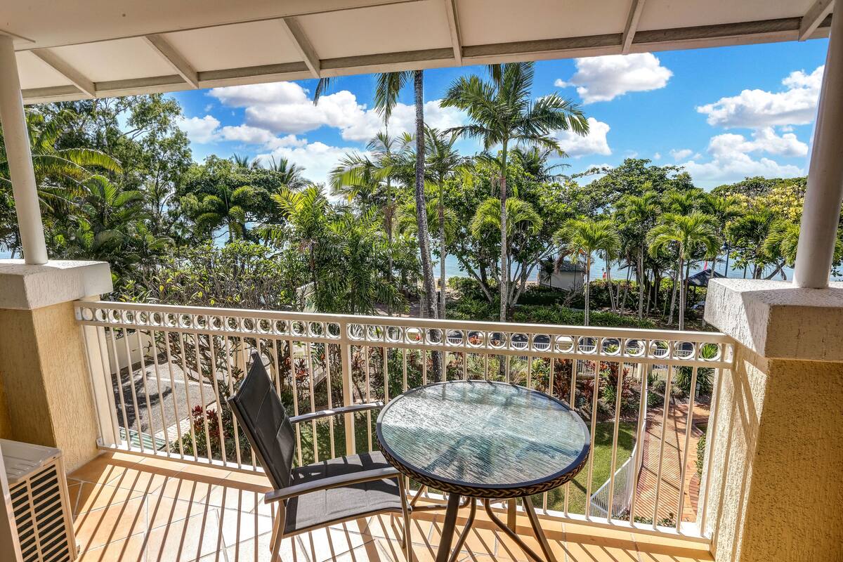 A balcony with a glass table and chairs, overlooking a tropical garden and a body of water.