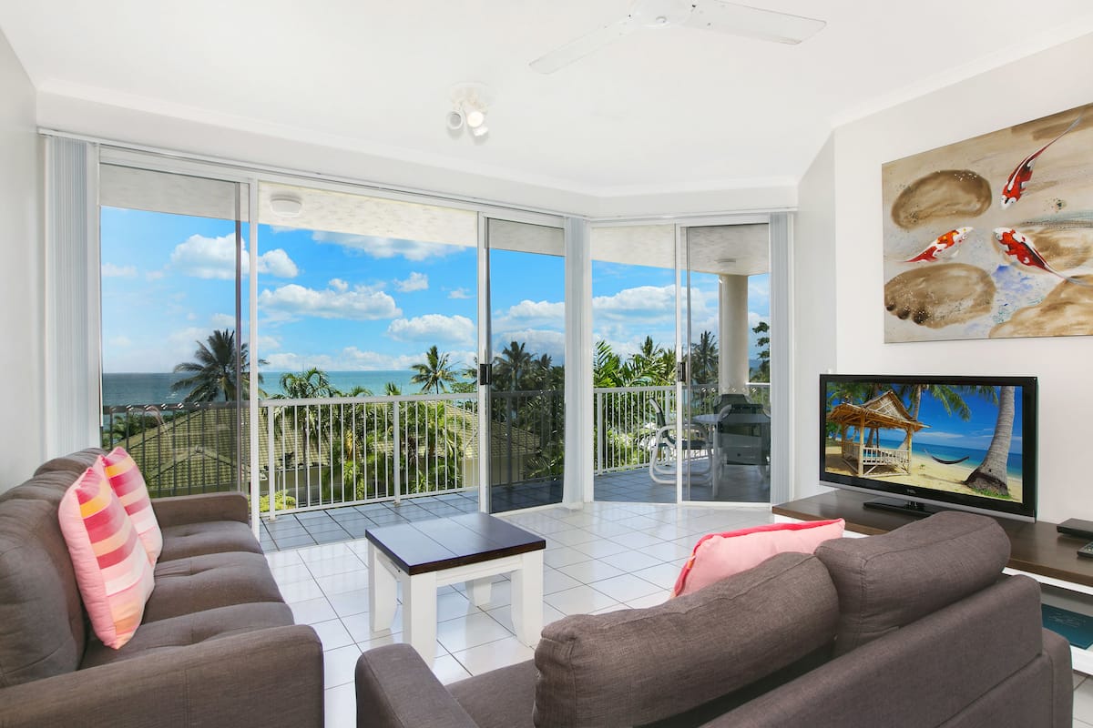 A modern living room with a large TV, grey sofas, a small table, and a balcony with a view of the ocean.
