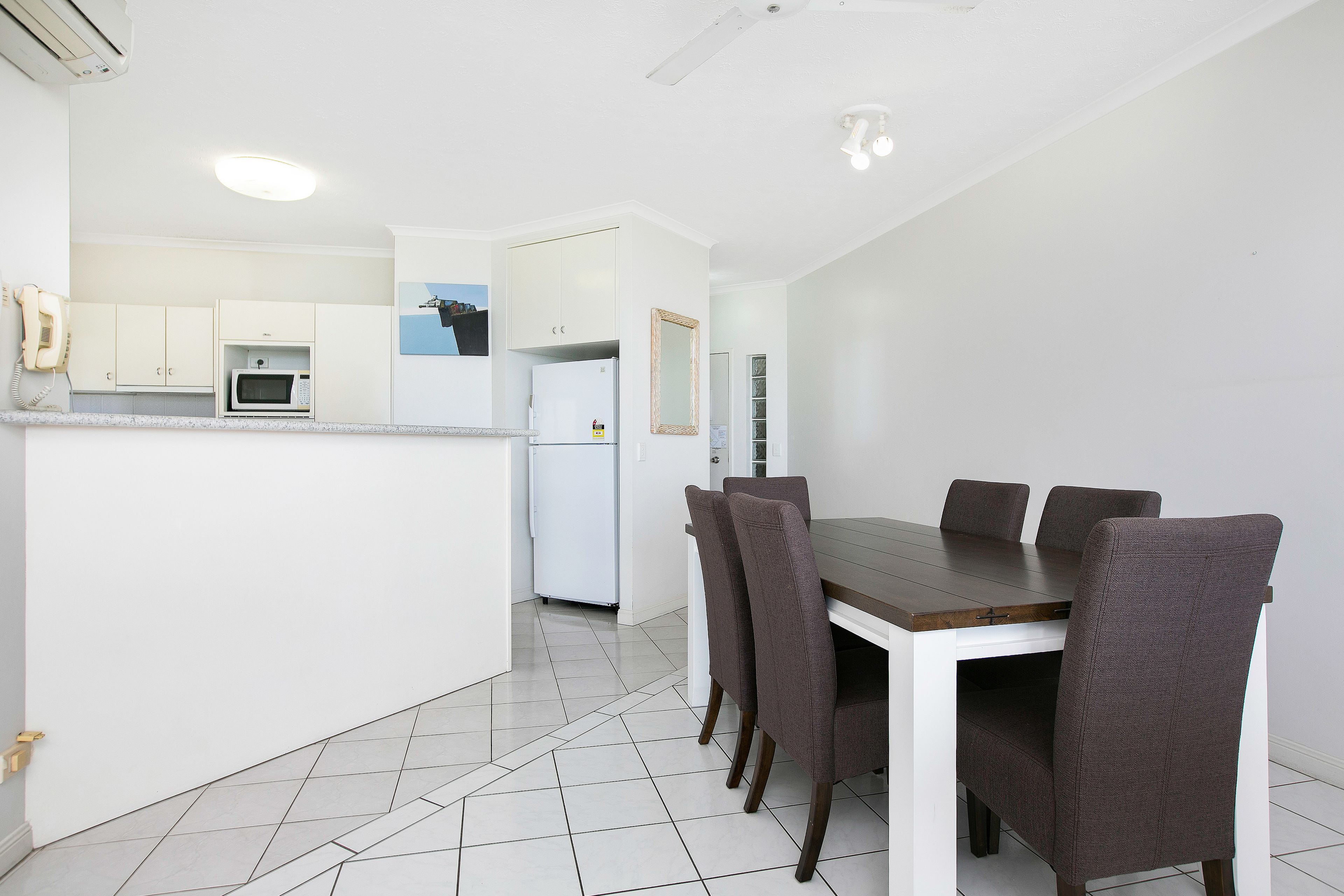 A modern kitchen with white cabinets, a microwave, and a refrigerator. There is a dining table with six chairs and a white floor.