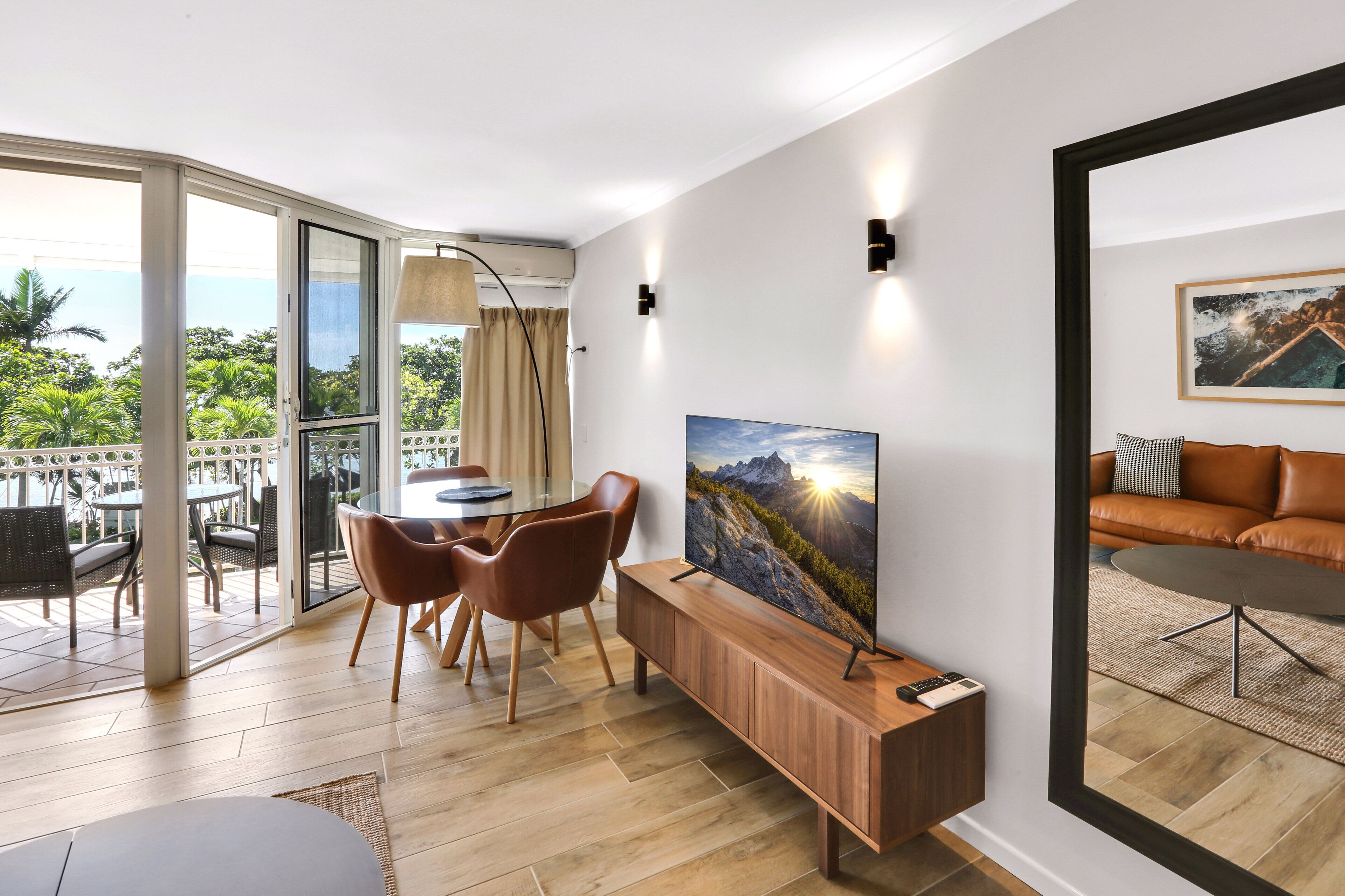 A modern living room with a large TV, wooden furniture, and a balcony with outdoor seating.