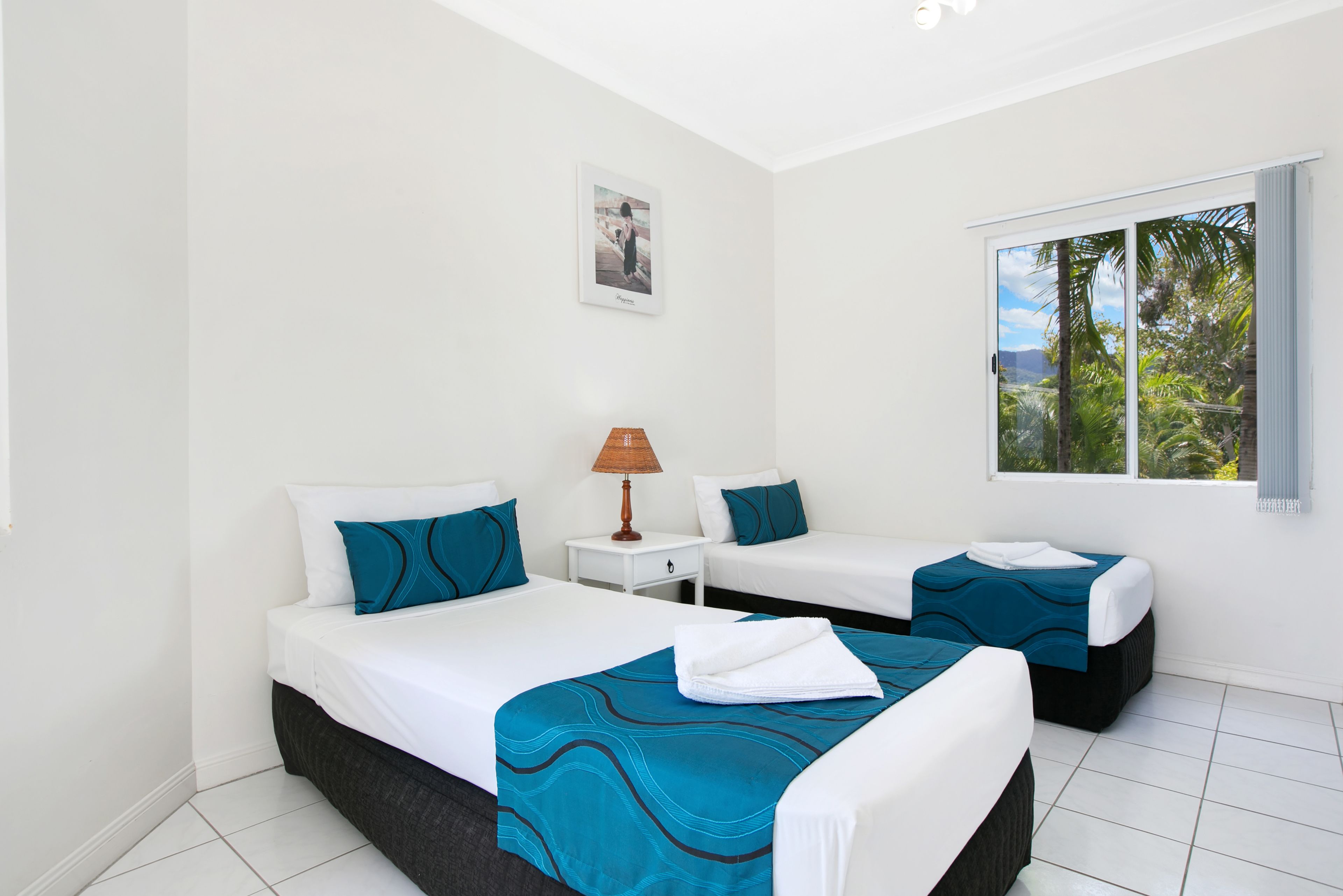 A hotel room with two beds, a nightstand with a lamp, a framed picture on the wall, and a window with a view of greenery.