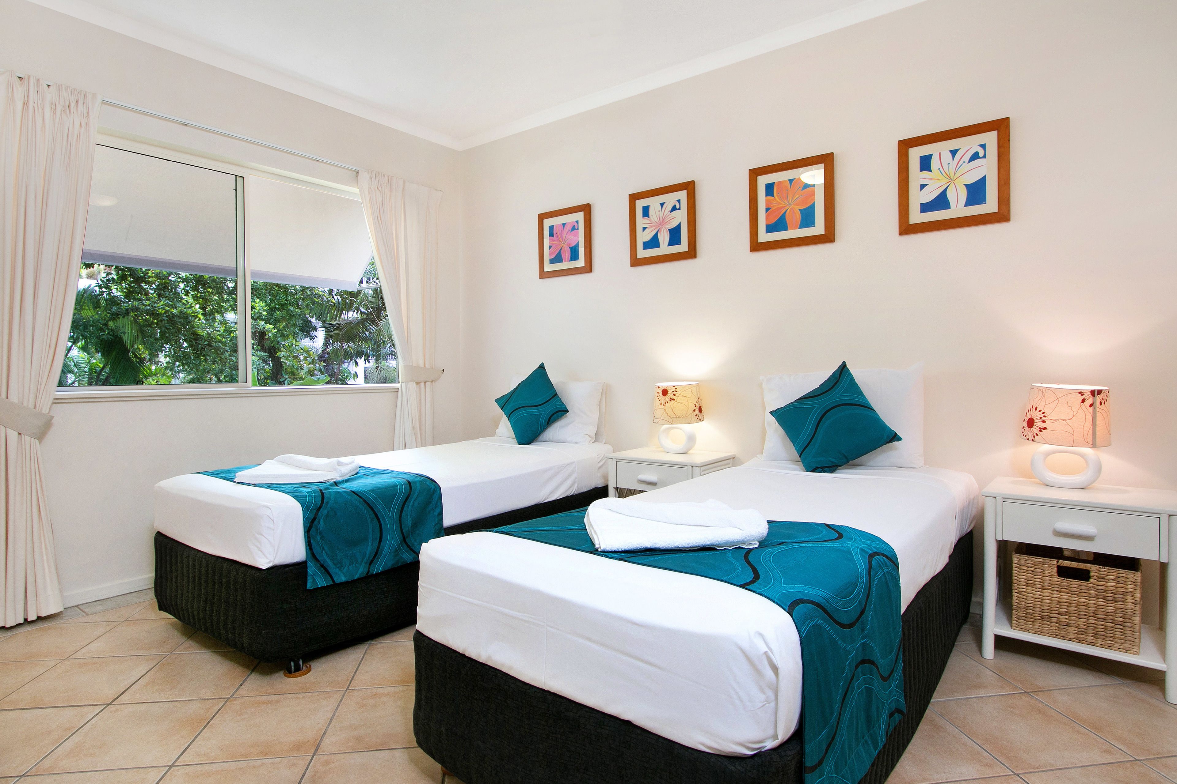 Habitación de hotel con dos camas, una ventana con cortinas, una mesita de noche con lámpara y decoración en la pared.