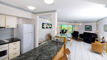 A modern kitchen with white cabinets, a black countertop, and a refrigerator. There is a dining area with a table and chairs, and a living room with a black sofa and a coffee table.