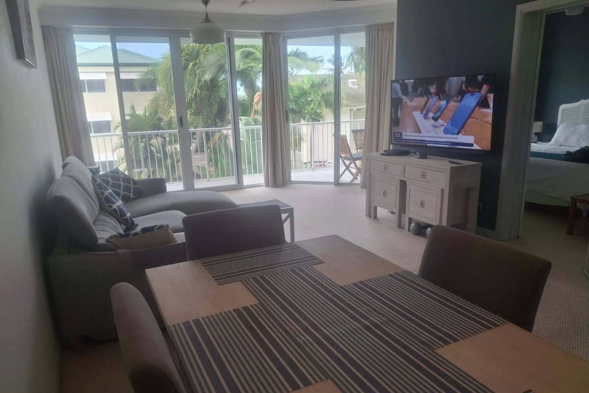 A living room with a sofa, a dining table, a TV, and a balcony with a view of palm trees.