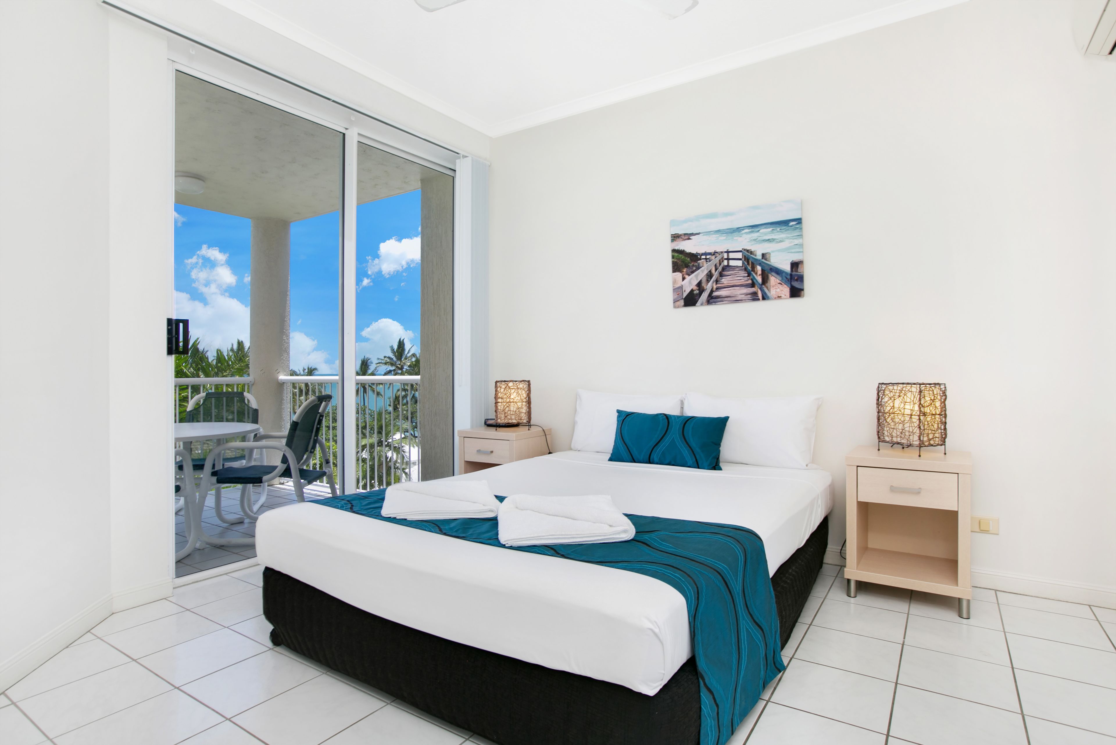 A bedroom with a large bed, a balcony with a table and chairs, and a painting of a beach scene.