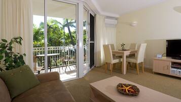 A living room with a sofa, a coffee table with a decorative bowl, a dining table with chairs, a TV on a cabinet, and a sliding glass door leading to a balcony with outdoor furniture and palm trees.