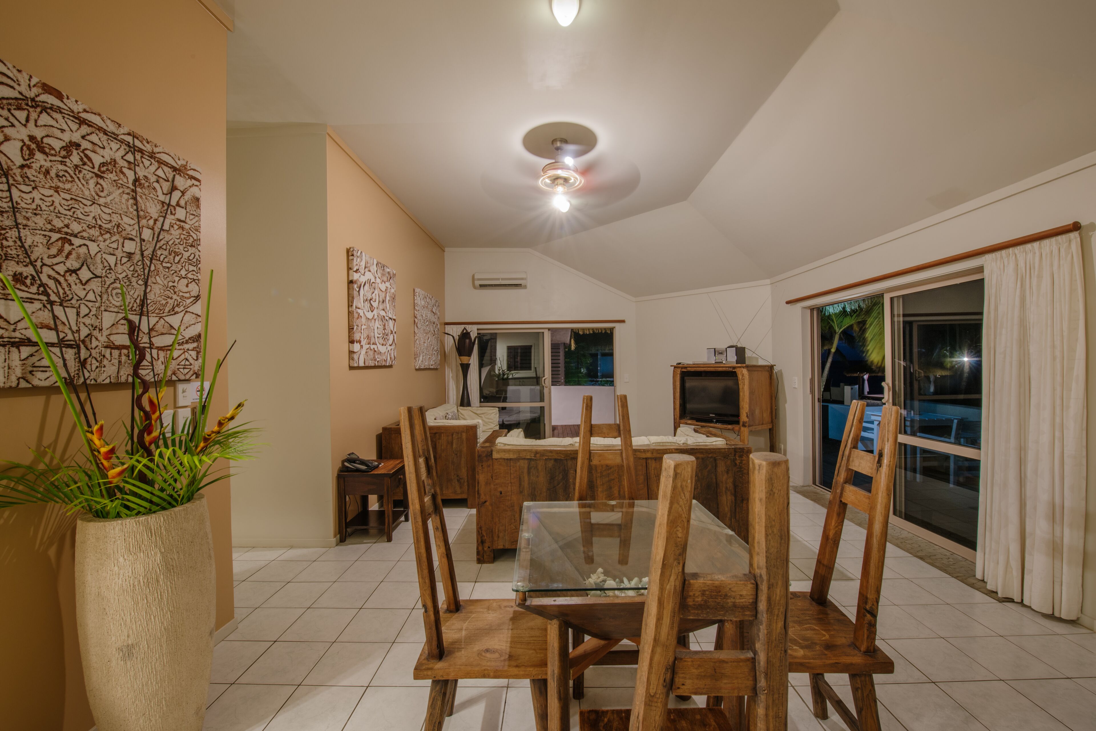 A dining area with a glass-top table and wooden chairs, a large vase with plants, and a ceiling fan.