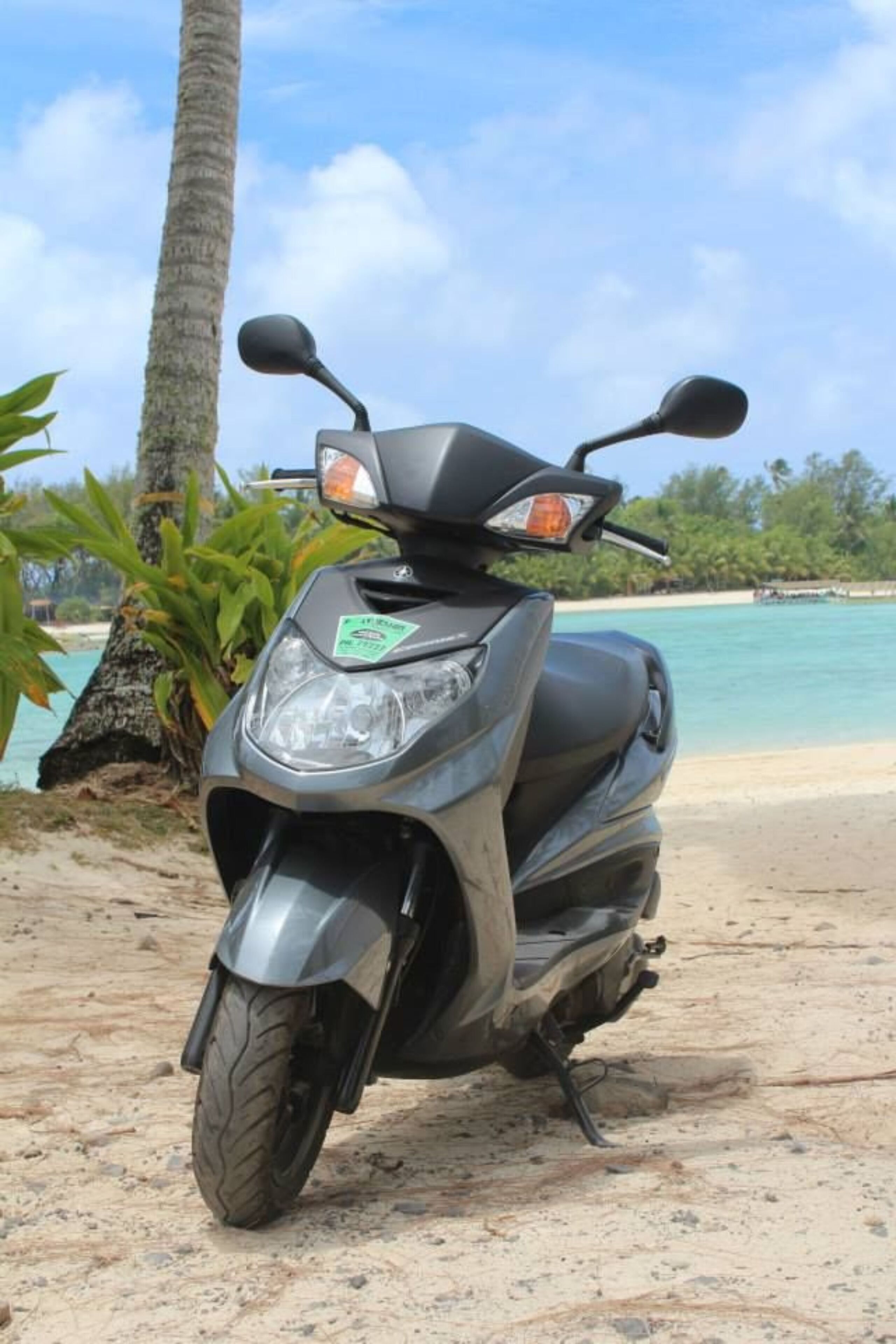 A scooter parked on a sandy beach with a palm tree and clear blue water in the background.