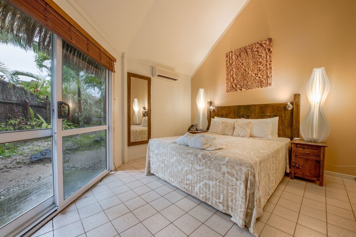 A bedroom with a large sliding glass door leading to a garden, a bed with a patterned bedspread, a wooden headboard, and a wall-mounted artwork.