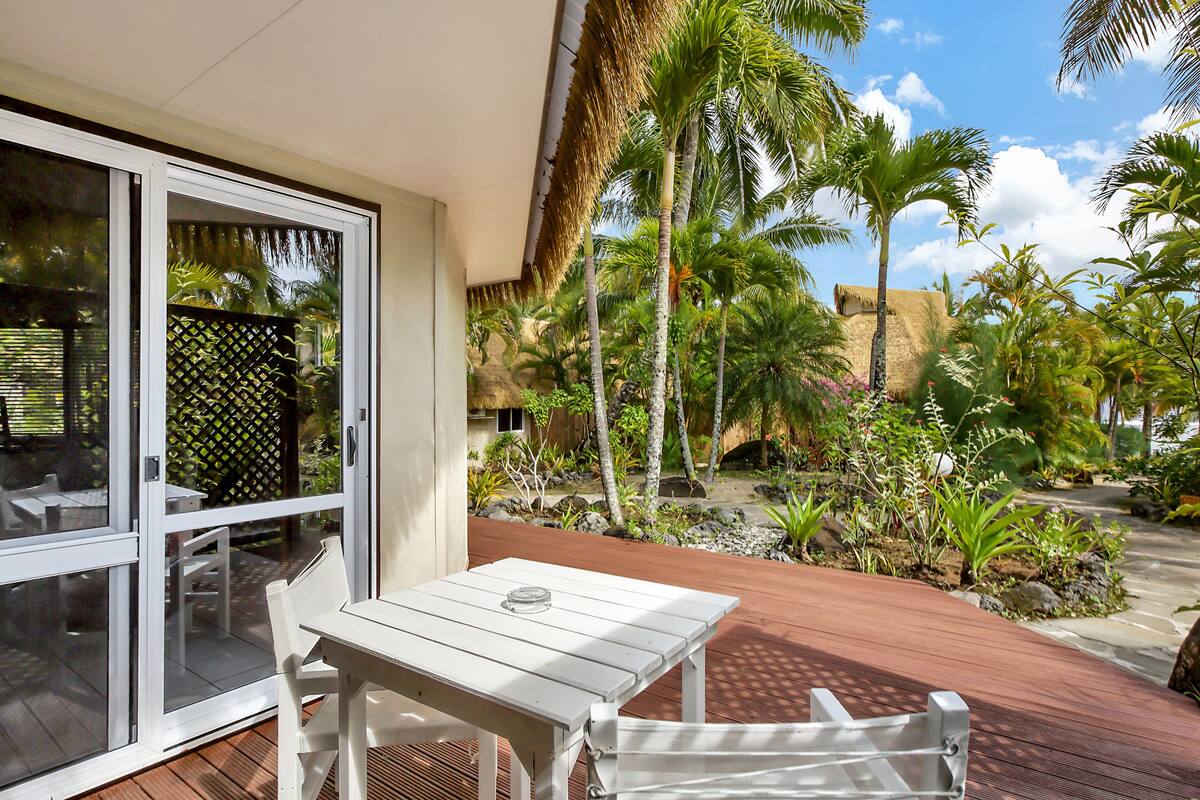 Un patio con mesa y sillas blancas, rodeado de exuberante vegetación tropical y una estructura de techo de paja.