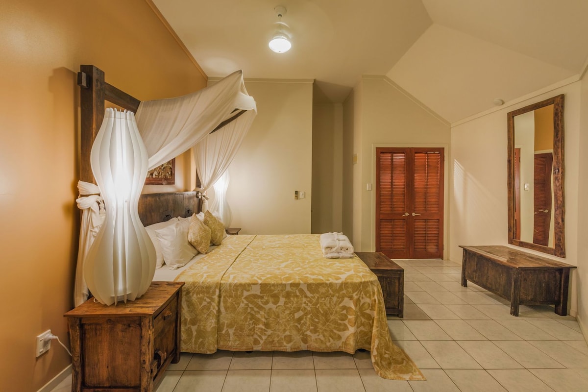 A bedroom with a canopy bed, a wooden bench, a mirror, and a wooden door.