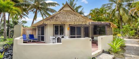 A thatched-roof bungalow with a patio, surrounded by tropical plants and palm trees.