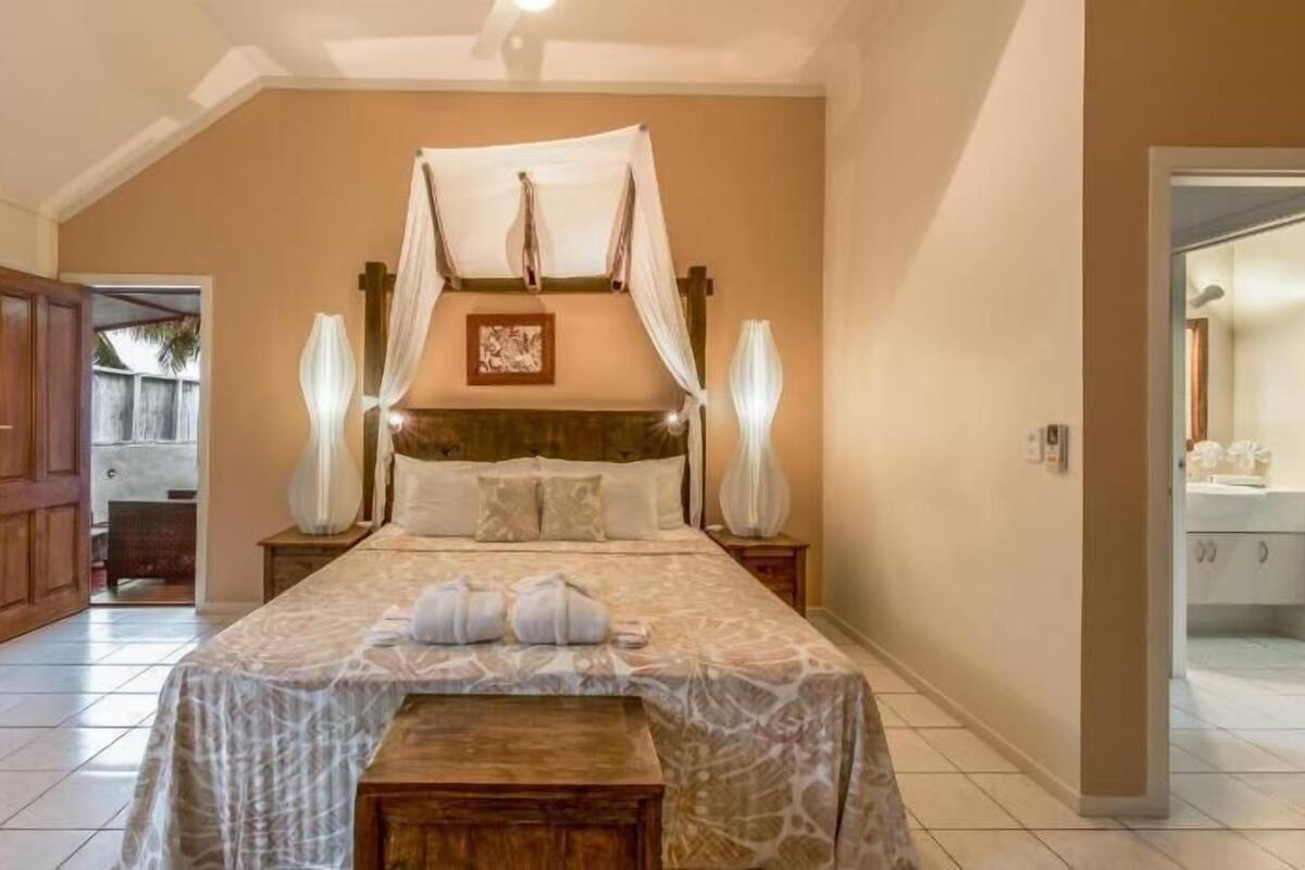 Un dormitorio con cama con dosel, un banco de madera, una mesita de noche y una mesita auxiliar con lámpara.