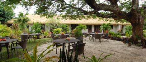 An outdoor dining area with tables and chairs, surrounded by plants and a large tree.