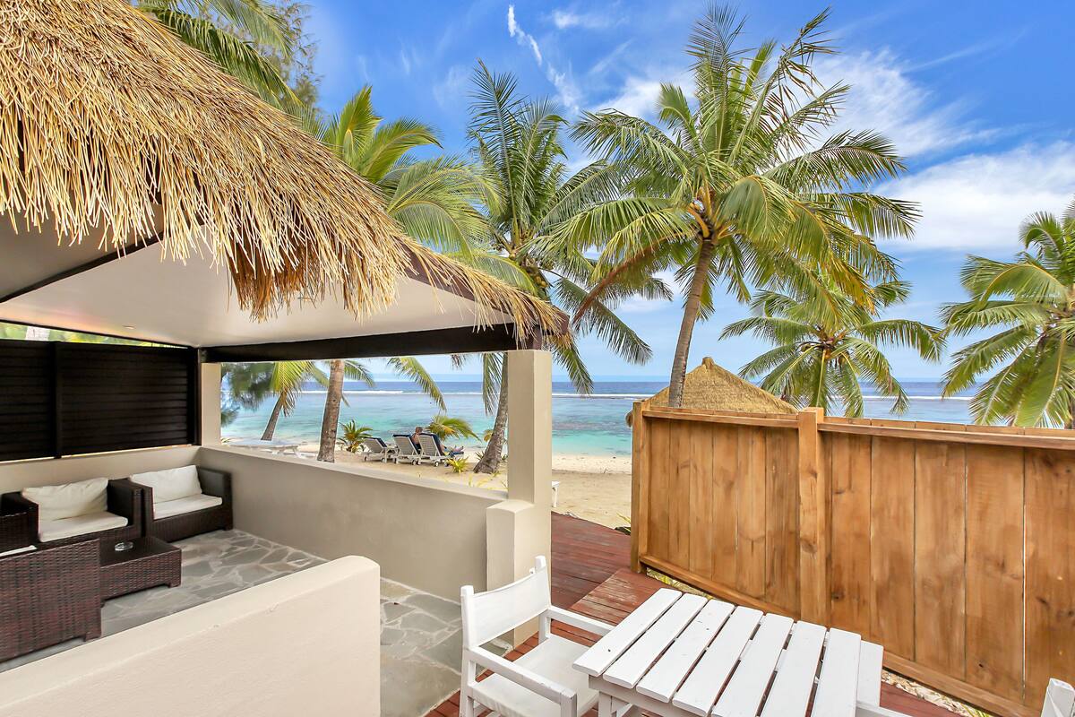 Un patio con techo de paja, mobiliario blanco y vistas a la playa y al océano.