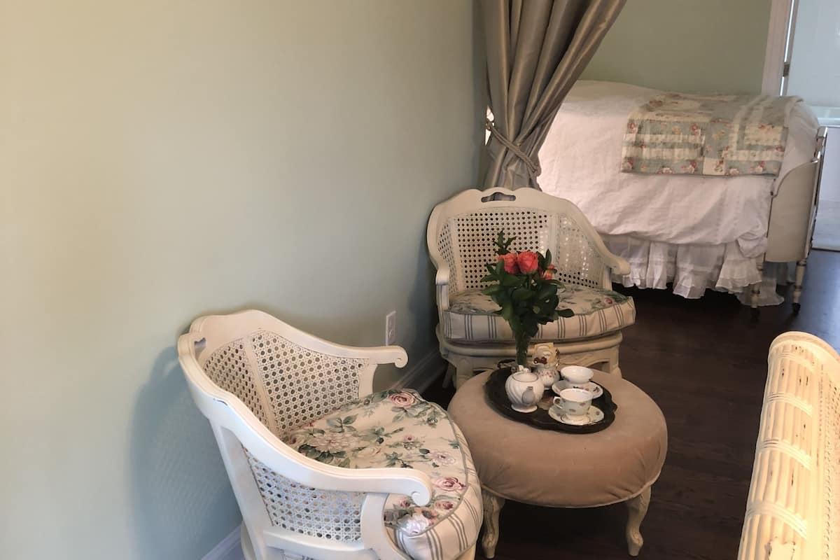 A room with two white wicker chairs, one with a floral cushion, and a small table with a teapot and cups. A bed with a floral quilt is in the background.