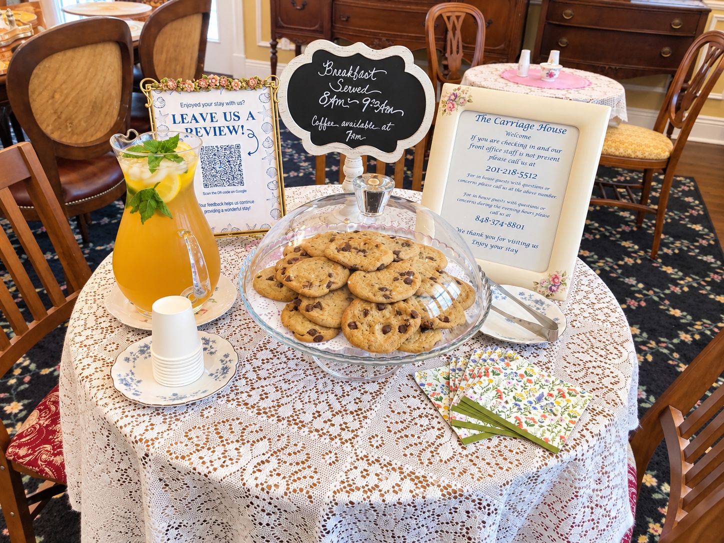 A table with a glass pitcher of iced tea, a bowl of cookies, a sign for breakfast service, and a welcome sign for The Carriage House.